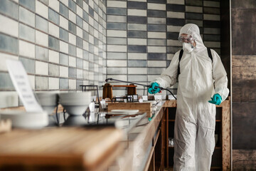 In a preventive isolated hotel dining room, a person in a special protective suit refreshes and cleans the air and space. He is spraying the counter of the buffet breakfast concept with alcohol. - obrazy, fototapety, plakaty