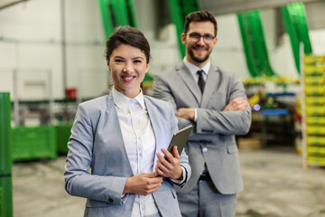 Naklejka na ściany i meble Business people in the warehouse. A man and a woman dressed in a fashionable suit stand in a warehouse and pose. A woman with a tablet in her hand is standing in front of a man with her arms crossed