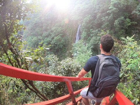 Back view of man with backpack standing on staircase with red metal railings in nature