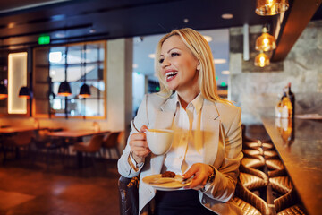 Confident and smiling blond woman elegantly dressed sitting at the bar in the hotel restaurant and having her coffee. Business lifestyle, taking a break, love my job