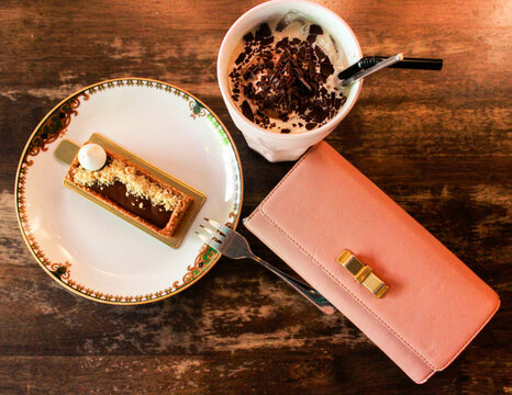 cake on plate and a cup of chocolate drink beside pink purse on wooden surface