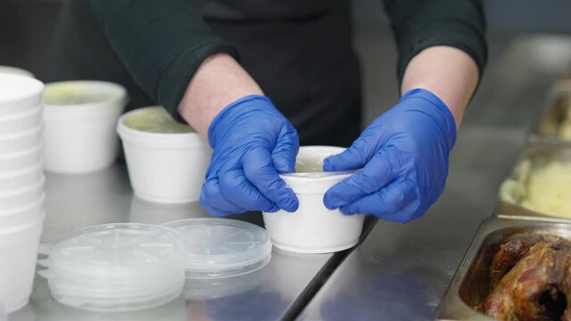 Female hands closing containers with hot soup for homeless people. Unrecognizable Caucasian woman in gloves working in commercial kitchen preparing food for impoverished beggar persons