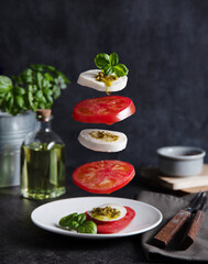 Concept levitation food. Italian caprese salad with tomatoes, mozzarella cheese, basil and pesto sauce flies over the plate on a dark background. Front view