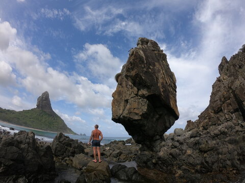 Man in swim suit standing beside a huge rock at the rocky beaches of Fernando de Noronha, Pernambuco, Brazil