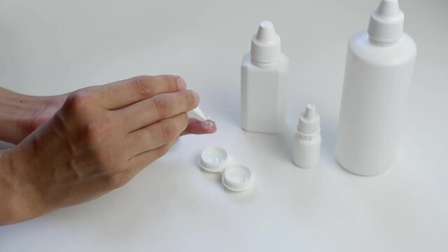A female hand with tweezers places a contact lens in a plastic container for storing lenses on a white background with vials, bottles with eye drops. Daily lens care.