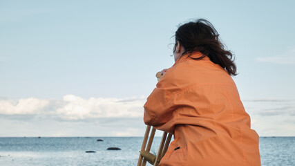 Fototapeta premium Woman sits on a stone leaning on crutches and looks at the lake