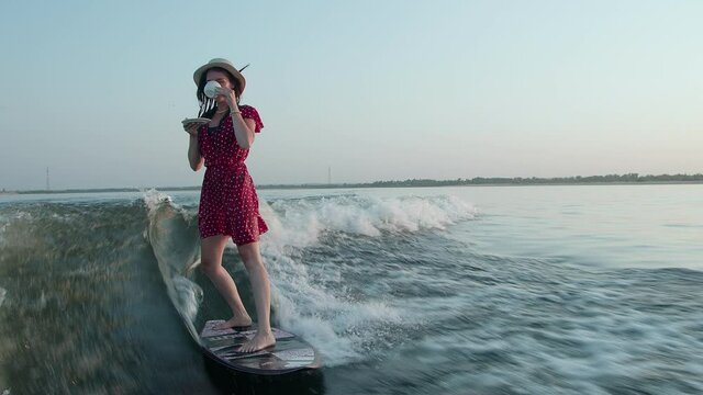 A surfer in a red dress and hat jumps on a wakeboard and holds a cup of tea or coffee in her hands. An experienced wakeboarder sprays water drops into the camera.