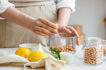 Woman squeezing lemon into blender with chickpeas on table in kitchen, closeup - obrazy, fototapety, plakaty