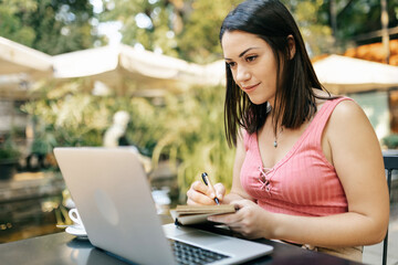 Millennial young hispanic woman working or studying on laptop while sitting at cafe - Freelancer businesswoman taking notes in a notebook while watching video call - Freelance people concept