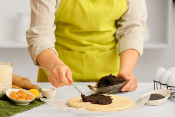 Woman preparing tasty poppy seed bun at table in kitchen, closeup
