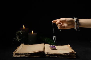 Woman holding pendulum above old book with gemstone against dark background