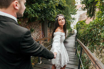 Beautiful bride and groom holding hands on wooden stairs surrounded by greenery in city.