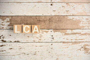 On a white damaged wood board, wooden word cubes are arranged in the letters LCA. It is an abbreviation for Life Cycle Assessment.