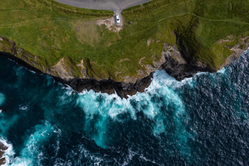 Birds eye view of car parked at end street of the famous Moher Cliff in Ireland with waves hitting the rocks during day