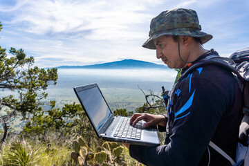 A portrait of a freelancer man with glasses with a laptop sitting on a rock in a mountain