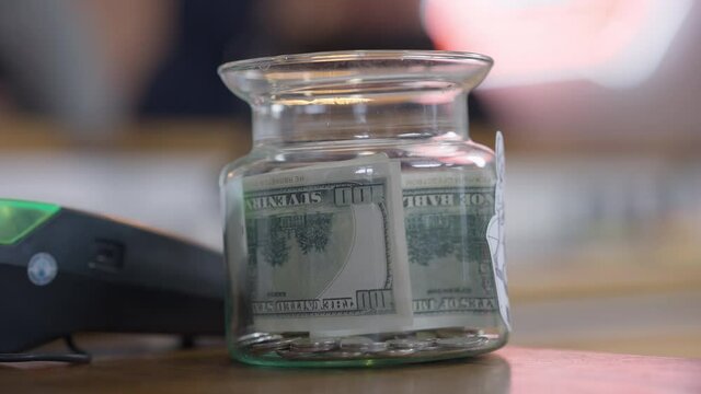 Close-up tips jar with female Caucasian hand putting cash inside. Unrecognizable cafe visitor leaving money for tips in cafeteria indoors