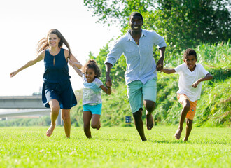 Cheerful mother, father, daughter and son gaily running in summer park during family walk