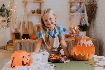 a blonde baby girl with long hair in an apron in the kitchen, decorated with pumpkins and garlands for Halloween, prepares a focaccia pie. space for text. High quality photo