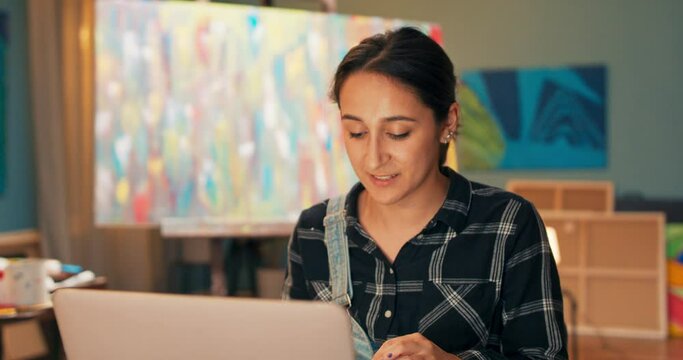 Young beautiful artist sits in studio in front of laptop screen, talks via webcam, social network with friend, describes her new painting, painting vision, shows found paints