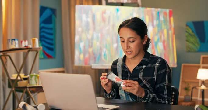 Portrait of art store employee, doing through computer, laptop live with store customers, woman showing new watercolor paints available for sale, advertising product, in background painting on easel