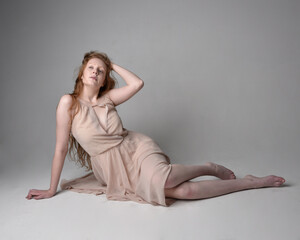 Naklejka premium Full length portrait of pretty red haired woman dancer, wearing skin toned flowing fairy dress. Sitting gestural poses isolated on studio background.