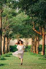 A beautiful Asian woman in a white dress walks along a pathway surrounded by trees.