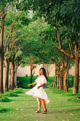 A beautiful Asian woman in a white dress walks along a pathway surrounded by trees.