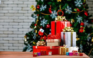 Colorful small and big present gift boxes with shiny ribbon bow tie placed on corner of wooden table in front fully decorative beautiful Christmas eve pine tree and brick wall in blurred background