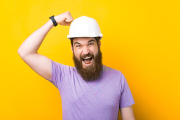 Excited bearded man is screaming at the camera while knocking in the hard hat he is wearing over yellow background.