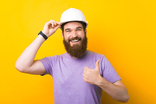 Wearing protection hard hat is a good idea - says bearded man who has one and shows thumb up over yellow background.