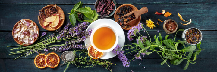 Tea panorama. Herbs, flowers and fruit around a cup of tea, an overhead flat lay shot on a dark rustic wooden background