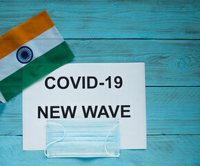 Fototapeta na wymiar On a blue wooden background lie a sheet with the inscription: covid -19 new wave, a medical mask and a flag of India