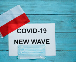 On a blue wooden background is a sheet with the inscription: covid -19 new wave, a medical mask and the flag of Poland