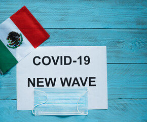 Fototapeta na wymiar On a blue wooden background are a sheet with the inscription: covid -19 new wave, a medical mask and a flag of Mexico