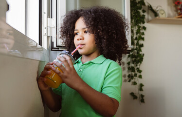 little afro boy drinks juice with straw at home at the window