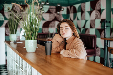 Stylish girl sits in a cafe and drinks coffee. Coffee to go in a cardboard cup. Woman with ginger hair in a beige warm suit in a cozy atmosphere. Modern interior. Calm and pleasant pastime