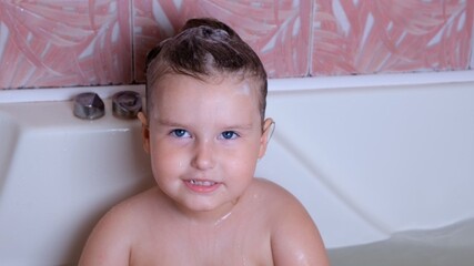 Little laughing girl 3 years old bathes and washes with foam on her head in water in the bathroom at home. Baby body care concept, hygiene.