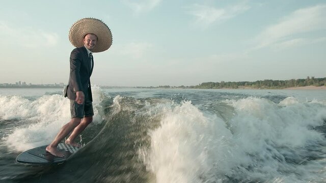 A surfer jumping on a wakeboard in a suit with a straw hat on his head. An experienced wakeboarder sprays water drops into the camera.