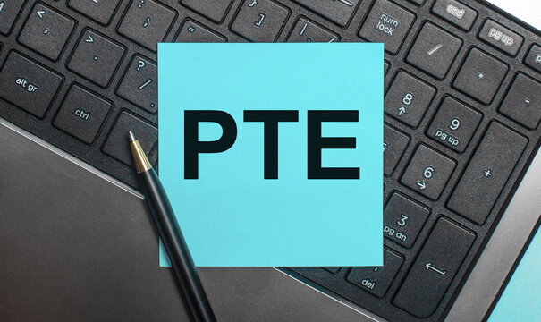 The computer keyboard has a pen and a blue sticker with the text PTE Pearson Test of English. Flat lay.