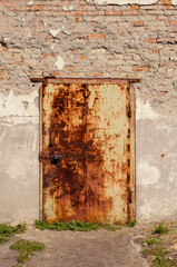 An old rusty metal door in a stone wall. The front door is locked with a padlock. Old red brick building. Plaster that has fallen off. Architecture.