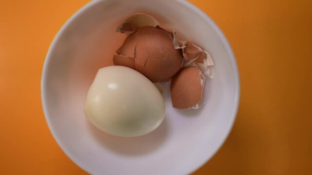 A boiled egg and egg shell in a ceramic bowl slowly moving on top of a orange background 