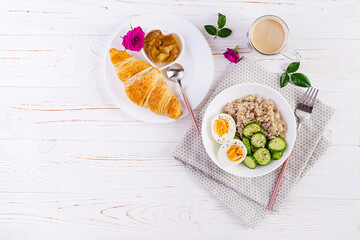 Breakfast oatmeal porridge with boiled egg, cucumber and croissant, jam, coffee. Healthy balanced food. Top view, flat lay