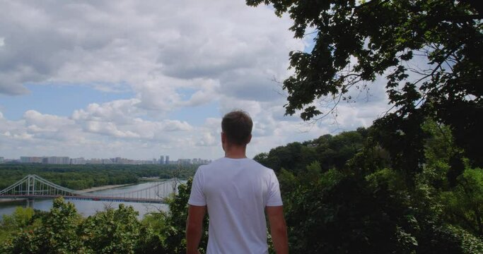 Rear view of young athletic man in white t-shirt stands on top of hill with river, bridge and city below. He raises his arms embracing the sky and feels like winner. Tracking shot, slow motion