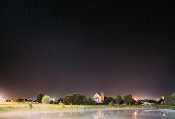 Night Sky Stars Over River Lake Near Houses In Village. Night Starry Sky Above Rural Landscape In Belarus. Glowing Stars Above Summer Nature