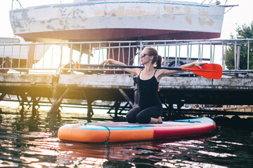 Close-up of a girl doing yoga on sub board. Psychology meditation, relaxation and self-healing concept. Lonely woman alone on board against a background of water and a pier. Stand up paddle boarding