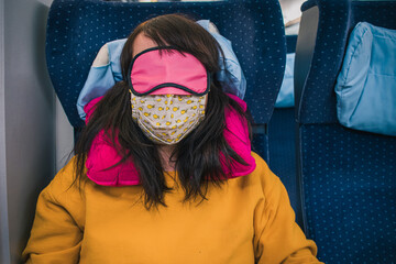 Young unknown woman with black hair is resting on a modern train wearing anti covid mask, eye mask and travel pillow. Colorful color combination. Sleepy woman on a train