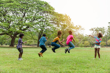Group of African American children having fun jumping over the rope in the park. Cheerful kid jumping over the rope outdoor. Happy black people enjoying playing together on green grass - obrazy, fototapety, plakaty