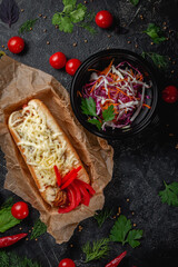 Delicious juicy hot dog with cheese sauce, tomato and fresh herbs in assortment, sandwich on the menu of a fast food restaurant on a dark stone table. Healthy option of fast food.