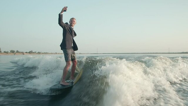 A surfer jumping on a wakeboard in a suit with a phone in his hands takes pictures of himself. A surfer takes a selfie and rides a wakeboard.