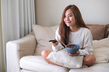 A young woman eating food while searching channel with remote control to watch tv on sofa at home